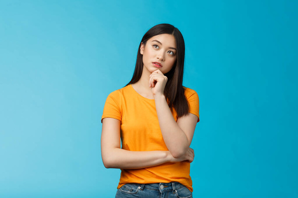 Thoughtful dreamy smart asian girl thinking, making plan inside head, touch chin, look up focused, contemplating interesting scene, pondering, making choice, taking decision, stand blue background - Foto, Imagen