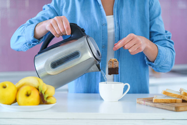 Woman putting tea bag in a cup and using an electric kettle for brewing hot tea at home at kitchen  - Foto, imagen