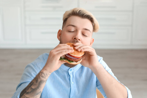 Young man eating vegan burger in kitchen - Фото, изображение