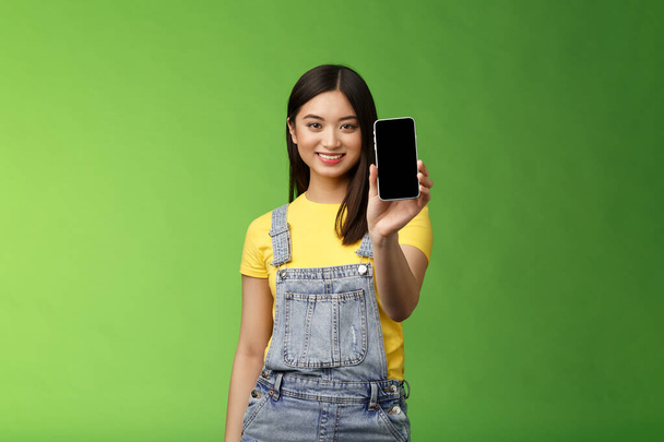 Впевнена в доброму погляді жінка-азіатка вводить додаток для смартфона, розширює руку тримає телефонну руку, посміхається в захваті, рекомендує соціальний профіль, хвалить власний блог, зелений фон
 - Фото, зображення
