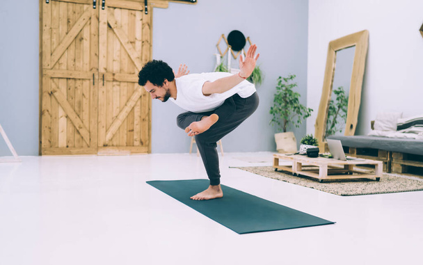Side view of man in t shirt and training pants doing yoga exercise in specific pose standing on mat on one leg - Fotografie, Obrázek