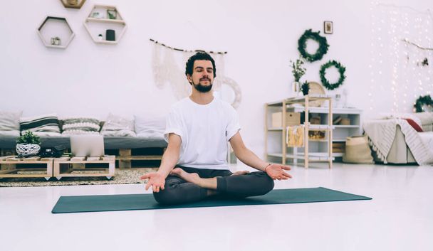 Calm bearded male with closed eyes sitting on sport mat and meditating in Padmasana in modern light living room at home - Zdjęcie, obraz