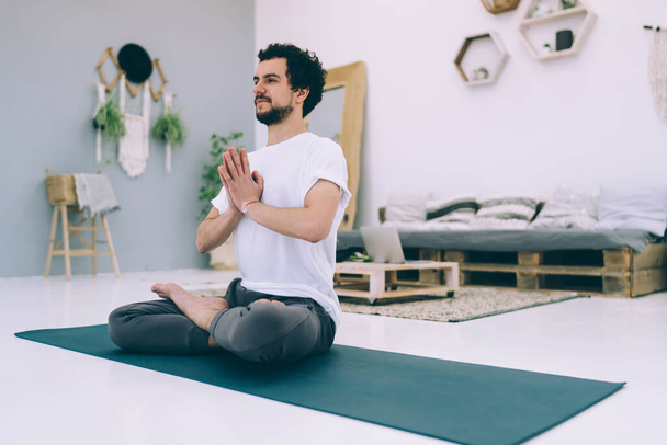 Side view of calm male looking away sitting on mat in lotus pose while practicing yoga in cozy room with decorative elements - Valokuva, kuva