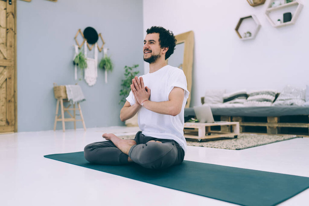 Side view of male with toothy smile and bare feet looking away while practicing Padmasana on sport mat in light living room - Foto, afbeelding