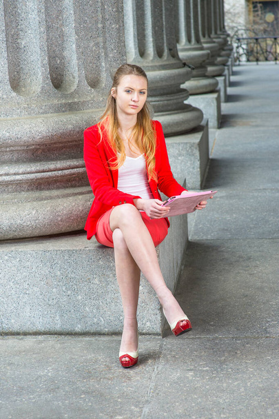 Habillée d'un blazer rouge, sous-vêtements blancs, jupe rouge et orteils ouverts talons hauts, jambes croisées, une jeune adolescente blonde est assise sur une base de colonne, étudiant sur une tablette - Photo, image