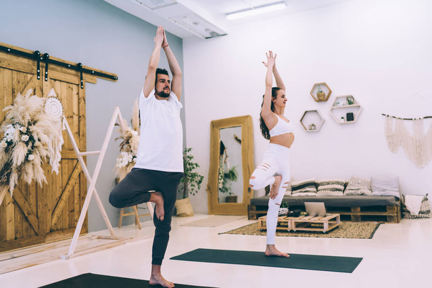 Full body of focused trainer with female in Ardha Padmasana with hands together above head strengthening muscles and improving balance in yoga studio - Valokuva, kuva