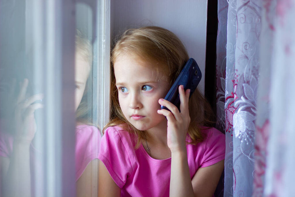 dziewczynka w różowej sukience mówi przez telefon siedząc przy oknie, nowe technologie - Zdjęcie, obraz