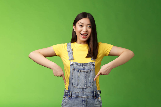 Живая энергичная азиатская милая женщина продвигает рекламу указывая пальцами вниз, показать вам прохладное место, улыбающаяся камера радостно, рассказывая большие приятные новости стенд зеленый фон эмоциональный - Фото, изображение