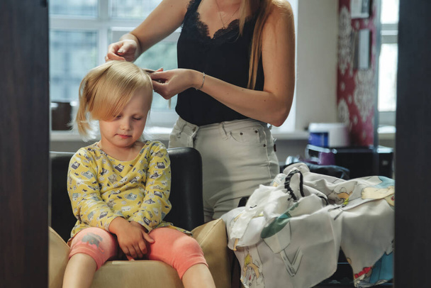 Barber woman make fashionable pretty hairstyle for cute little blond girl child in modern barbershop, hair salon. Hairdresser makes hairdo for young baby in barber shop. Concept hairstyle and beauty - Foto, immagini