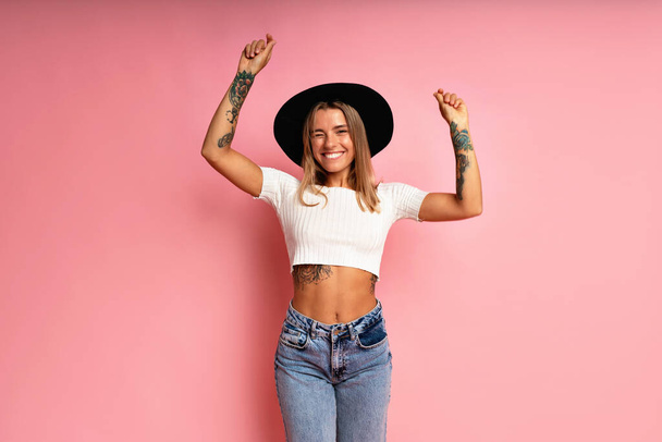 Hipster stylish girl posing at pink background, withe crop top jeans and black fedora playful happy mood. - Foto, Imagem
