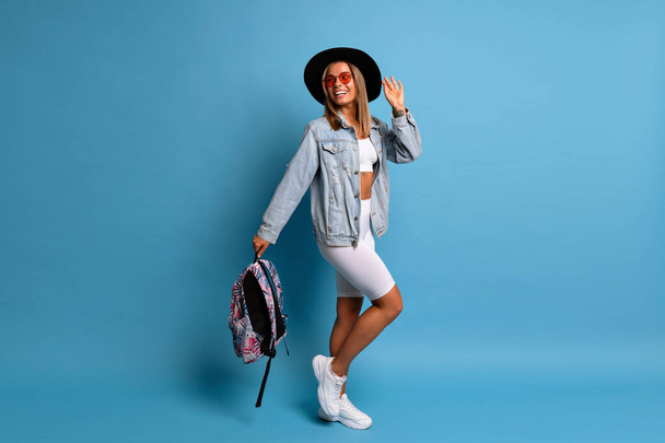Trendy fit hipster blonde own posing at studio blue background, full lenght, copy space. Trendy casual tourist outfit. - Foto, imagen