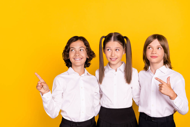Photo of three schoolkids direct finger empty space look up wear white shirt isolated yellow color background - Foto, imagen