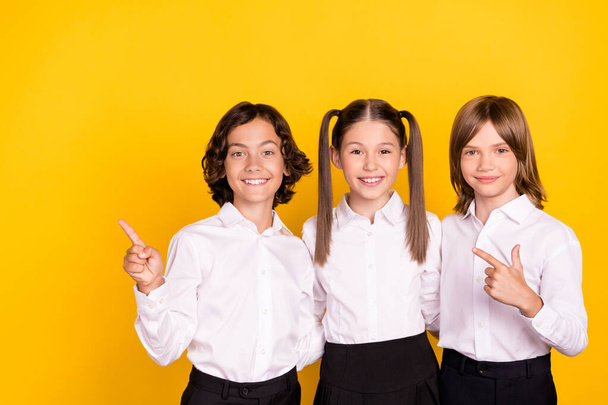 Photo of three schoolkids indicate finger empty space promote sale wear white shirt isolated yellow color background - Фото, изображение