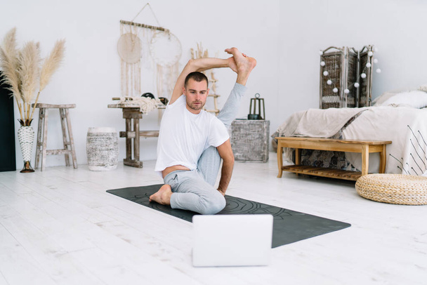 Serious concentrated male in casual wear sitting in asana training online via laptop computer at home interior, calm caucasian hipster guy enjoying yoga practice video lessons doing poses at apartment - Foto, immagini