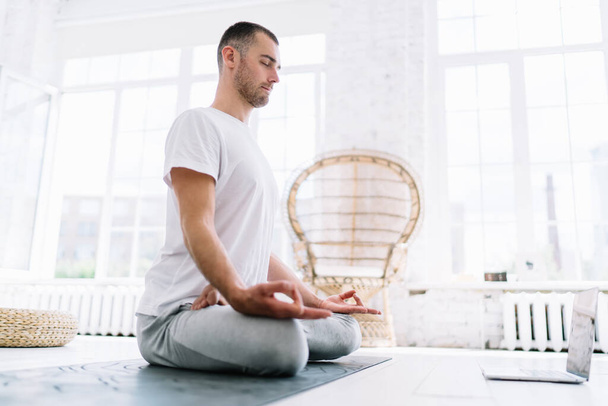 Calm caucasian man with closed eyes sitting in lotus pose meditating feeling mindfulness and spirituality, handoms hipster guy using laptop computer for training yoga online staying on isolation - Foto, Imagem