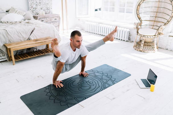 Concentrated strong man in casual wear having morning training for keeping fit using laptop computer watching video yoga practice,serious caucasian hipster guy standing in asana breathing and rest - 写真・画像