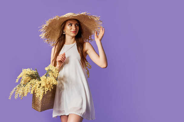 Happy woman in summer white dress, straw hat with bunch of flowers in bag posing on purple, velvet studio background - Photo, image