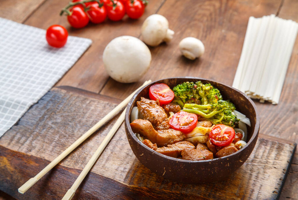 Udon with chicken in Japanese sauce on a plate of coconut shells on a stand next to vegetables. Horizontal photo - Fotografie, Obrázek