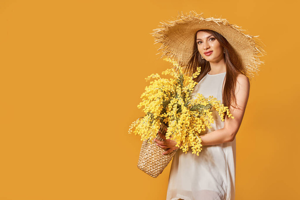 Happy woman in summer white dress, straw hat with bunch of flowers in bag posing on yellow studio background - Фото, зображення
