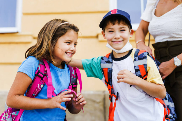 Kleine Mädchen und Jungen, kaukasische Geschwister oder kleine Freunde umarmen sich mit Rucksäcken auf den Schultern auf dem Rückweg zur Schule - Kleine Erstklässler im Sommertag lächeln selektiv - Foto, Bild