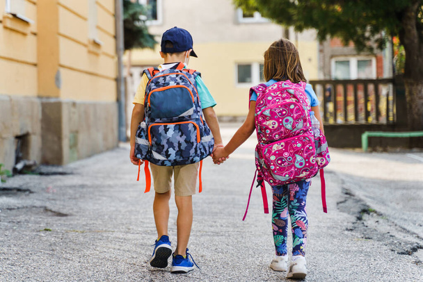 Back view full length portrait of small caucasian boy and girl brother and sister or little friends children leaving school first grade pupils with backpack bags on their back in sunny day outdoor - Photo, Image
