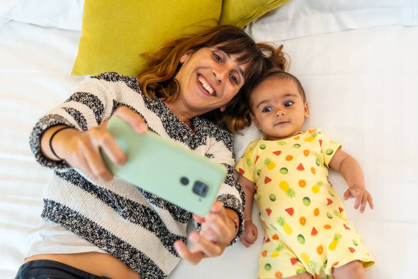 Young mother with her baby taking a selfie in her bedroom bed, first-time Caucasian with her four-month-old baby, family at home - Fotó, kép