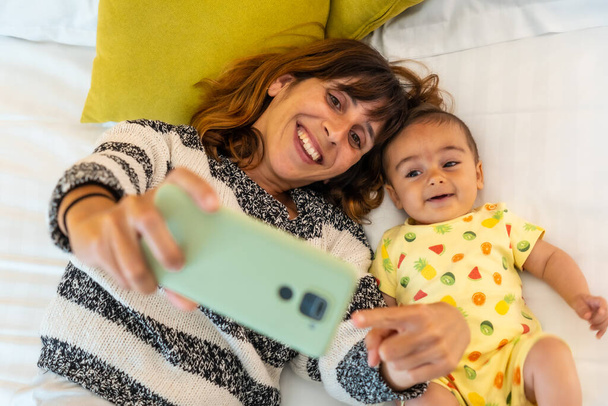 Young mother with her baby taking a selfie with the mobile phone on the bed in her bedroom, family at home - Fotoğraf, Görsel