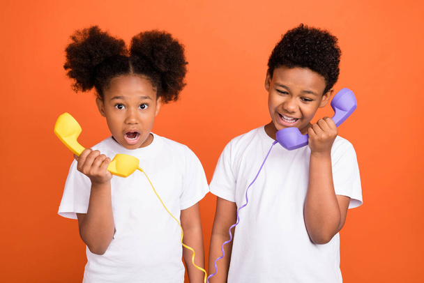 Zdjęcie małego pod wrażeniem dziewczyna chłopiec rozmawiać telefon nosić biały t-shirt izolowane na pomarańczowym tle - Zdjęcie, obraz