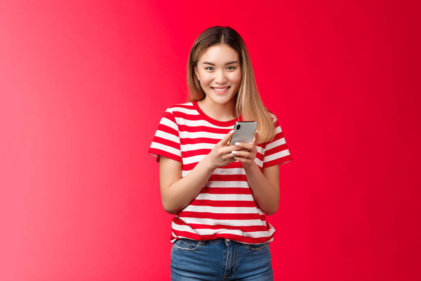 Iloinen kaunis aasialainen vaalea tyttö kikattaa hymyilevä hammas, onnellinen, tilalla älypuhelin lukea ystävä hauska viesti, seistä punainen tausta rennosti, pyyhkäisemällä dating sovellus, ostoksia verkkokaupasta huoleton - Valokuva, kuva