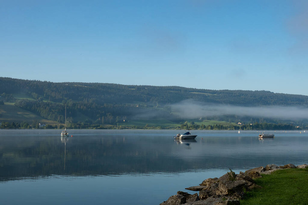 Varhain aamulla Lac de Joux, Sveitsi: Misty Jura kukkulat järven takana makaa veneitä  - Valokuva, kuva