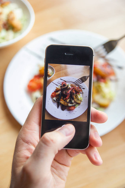 Ένας άντρας φωτογραφίζει μια νόστιμη σαλάτα στο κινητό του σε ένα εστιατόριο. - Φωτογραφία, εικόνα