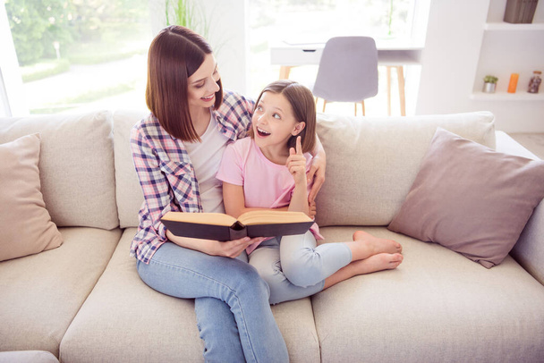 Portrait of two attractive cheerful girls sitting on divan reading book hugging at home flat indoors - Fotoğraf, Görsel