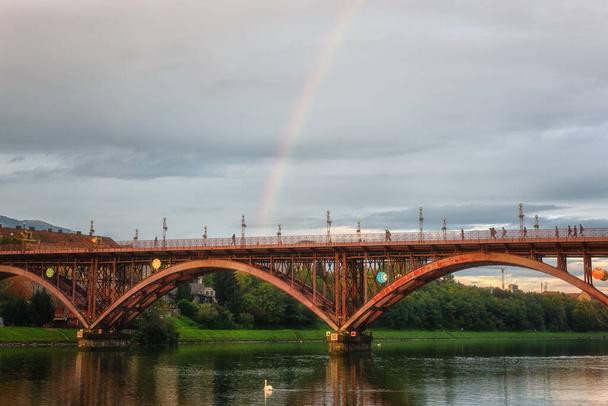 Ponte velha (Stari mais) ou ponte principal (Glavni mais) através do rio Drava, Maribor, Eslovênia. Paisagem urbana panorâmica com construção metálica, ribeira verde e céu nublado com arco-íris - Foto, Imagem
