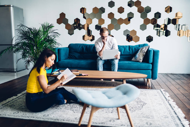 Young bearded man and African American woman in comfort outfit sitting at living room with laptop and notebook and creating business plan for startup - Фото, изображение