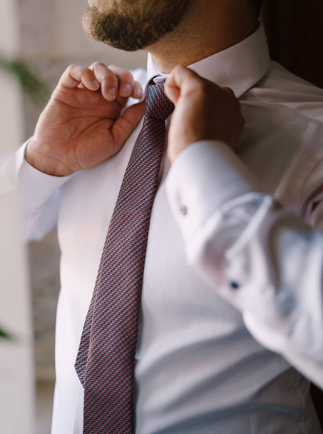 Ο γαμπρός σφίγγει μια πολύχρωμη γραβάτα σε ένα λευκό πουκάμισο. Κοντινό πλάνο - Φωτογραφία, εικόνα