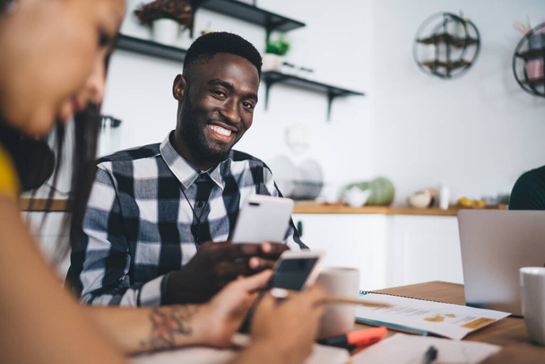 Young black student in casual clothes with mobile phone smiling at camera while sitting at kitchen table with crop fellow students using gadgets - Foto, Imagen