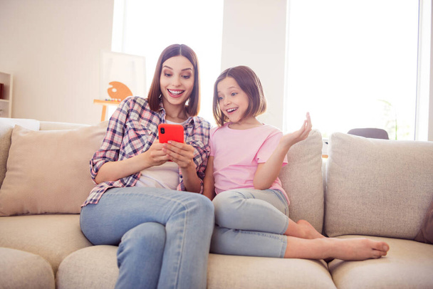 Portrait of two attractive cheerful girls sitting on divan using device gadget having fun at home flat indoors - Photo, image