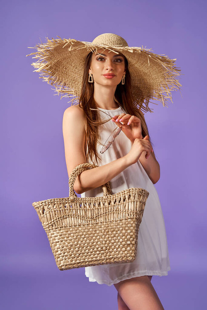 Beautiful young woman in white dress, summer straw hat, bag isolated on pastel violet background.  - Foto, immagini