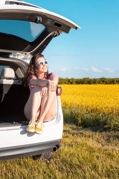 smiling pretty woman sitting in suv car trunk on sunset looking at farm field road trip concept - Foto, Imagen