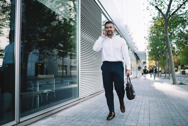 Middle aged businessman with trendy briefcase in hand walking at urban street using cellphone for making call and discuss work, confident male entrepreneur connecting to roaming for mobile talking - Foto, Imagem