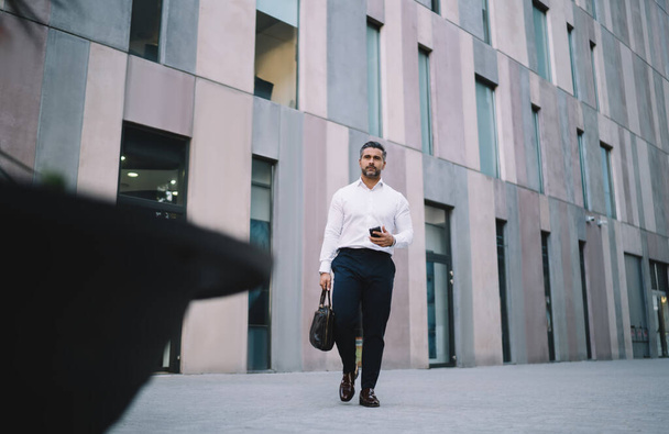 Mature businessman dressed in formal wear walking at downtown urbanity and using cellphone gadget, middle aged banker with trendy briefcase and cellphone in hands walking in financial district - 写真・画像