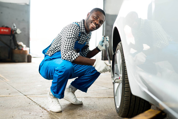 Happy african mechanic with ratchet wrench repairing tire in white gloves and blue dickey. Replacing tire - Fotoğraf, Görsel