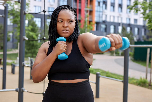 Serious overweight fat african woman exercising with dumbbells, outdoor sport activity - Photo, image