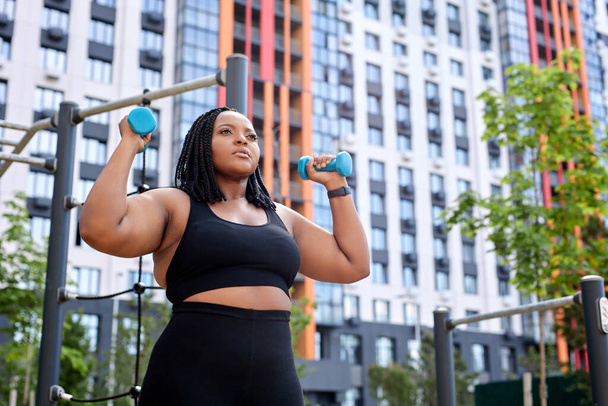 Overweight african woman holding two blue dumbells. Healthy life. Fitness concept - Фото, зображення