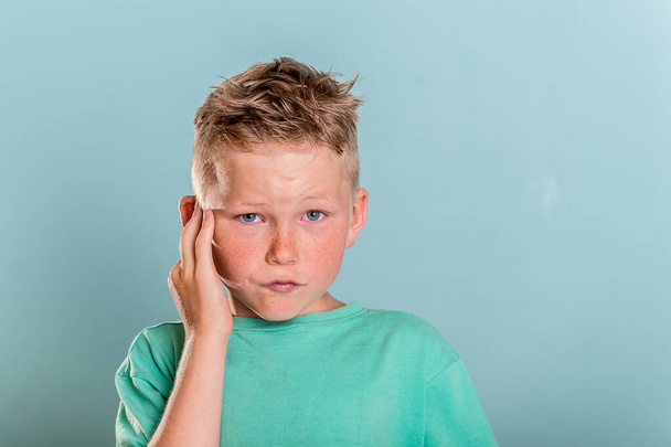 Chlapec se dotýká svého bolestivého ucha izolovaného na modrém pozadí. Škola se dívá do kamery s rukou na uchu a žádá o pomoc. koncepce zdravotní péče. Blonďatý portrét dítěte - Fotografie, Obrázek