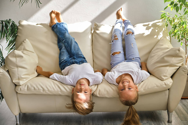 Hezká holčička a chlapec leží na zádech na pohovce v obývacím pokoji, dívají se do kamery a usmívají se, zatímco si hrají doma - Fotografie, Obrázek