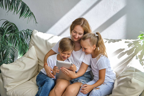 Młoda matka z synkiem i córką bawią się cyfrowym tabletem na kanapie w domu - Zdjęcie, obraz