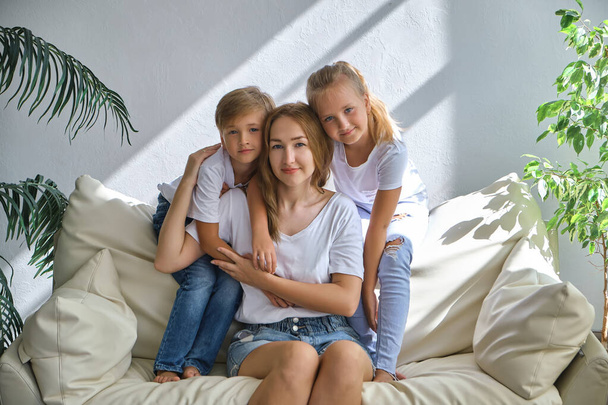 Adorable Little kids, boy and girl hug their mom while sitting on the sofa in the living room on a sunny day - Фото, изображение