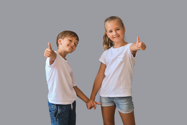 Cute two kids, little boy and girl in white t-shirts show thumbs in air recommending, hold hands best friends isolated on gray background - Photo, image