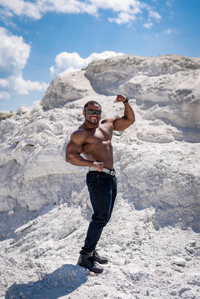 Vertical view of the strong fit bodybuilder showing his biceps. Handsome athletic man showing his muscles while standing at the sand with blue sky at the background - Foto, afbeelding